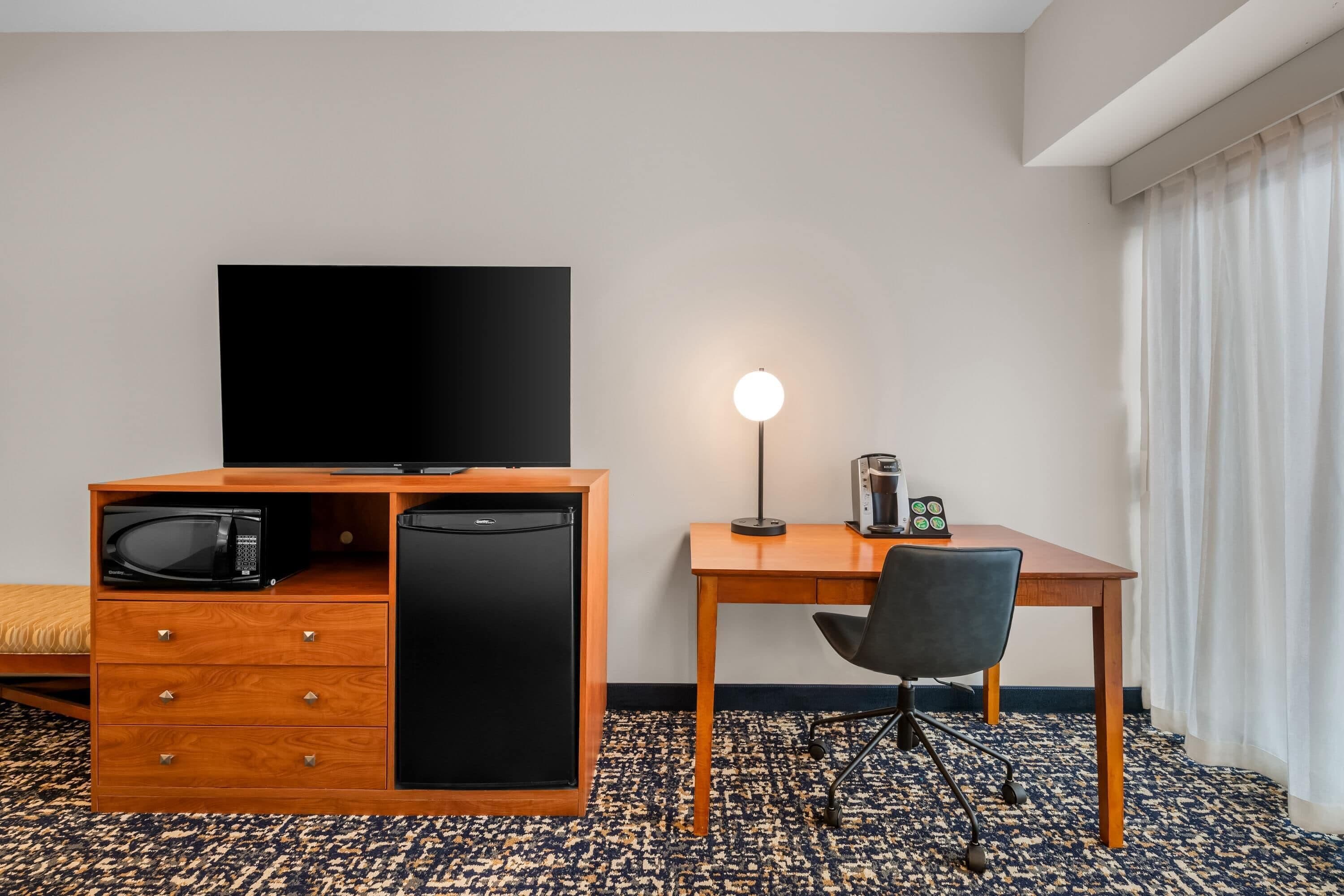 A hotel room with a wooden desk, a black television, a microwave, a refrigerator, a lamp, and a chair.
