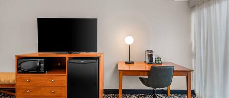 A hotel room with a wooden desk, a black television, a microwave, a refrigerator, a lamp, and a chair.