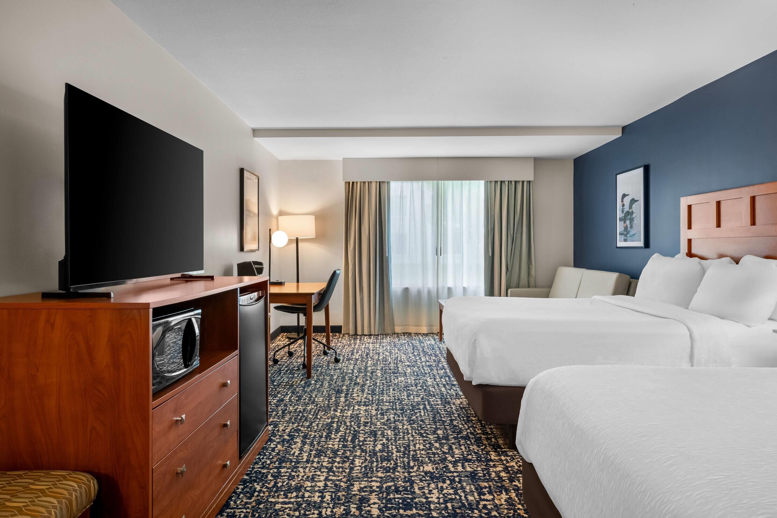 A hotel room with a TV, desk, two beds, a window with curtains, and a framed picture on the wall.