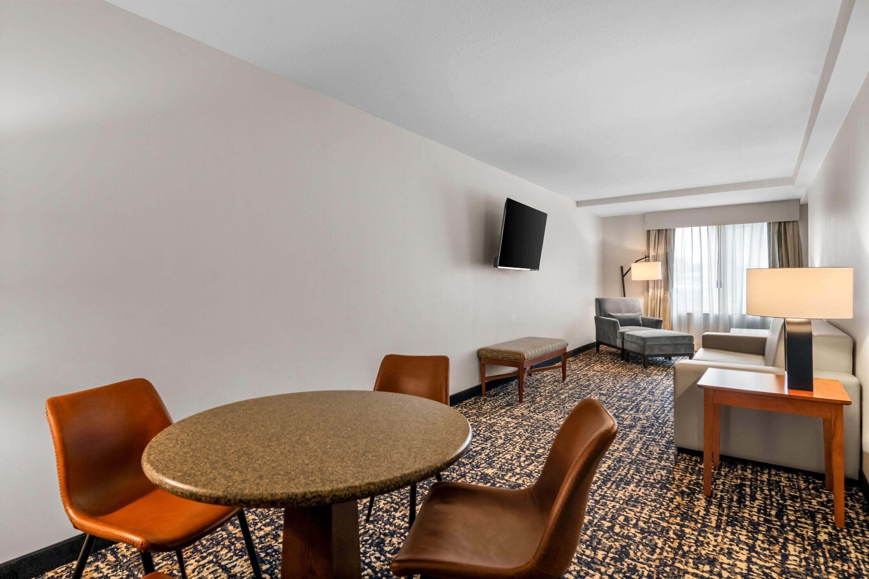 A hotel room with a round table, four chairs, a flat-screen TV mounted on the wall, a sofa, a lamp, and a patterned carpet.