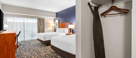 A hotel room with two beds, a desk, a chair, a hanging coat, and a window with curtains.