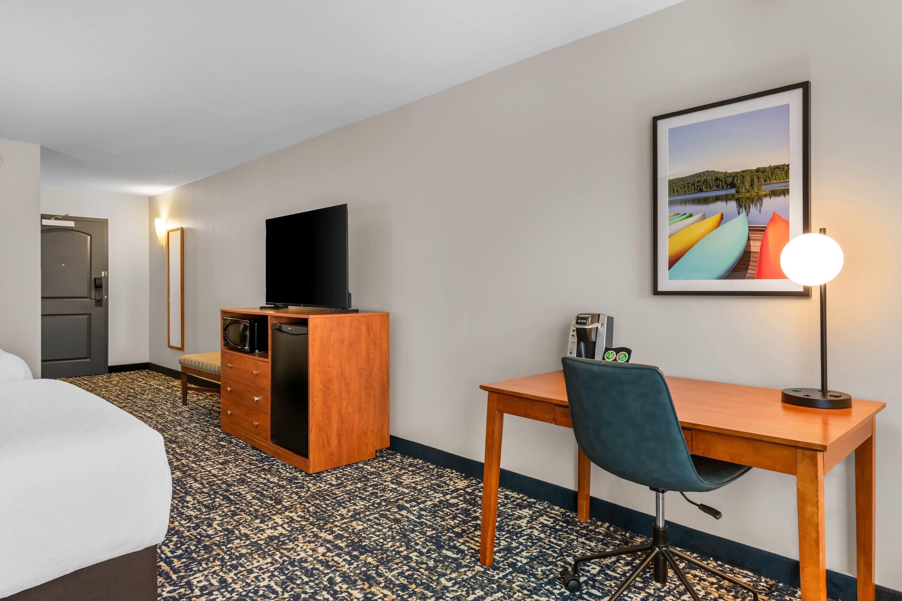 A hotel room with a bed, a desk with a chair, a television on a wooden cabinet, a framed picture on the wall, and a lamp.