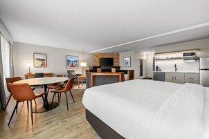 Habitación de hotel moderna con una cama grande, una mesa de comedor con sillas, una pequeña cocina y obras de arte en las paredes.