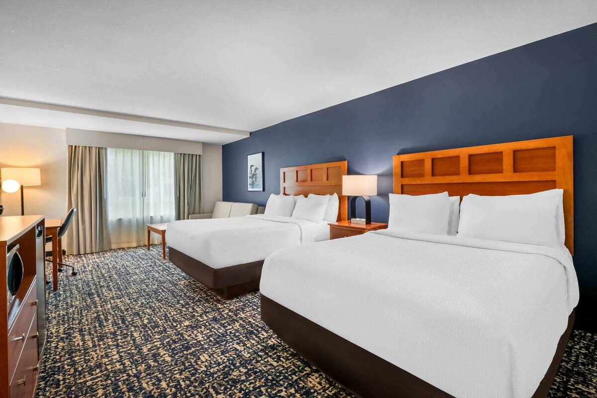 A hotel room with two beds, a desk, a chair, a lamp, and a view of the outdoors.