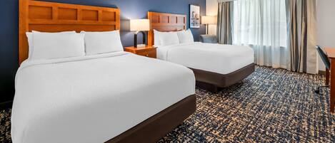 A hotel room with two beds, a large window with curtains, a desk, and a patterned carpet.
