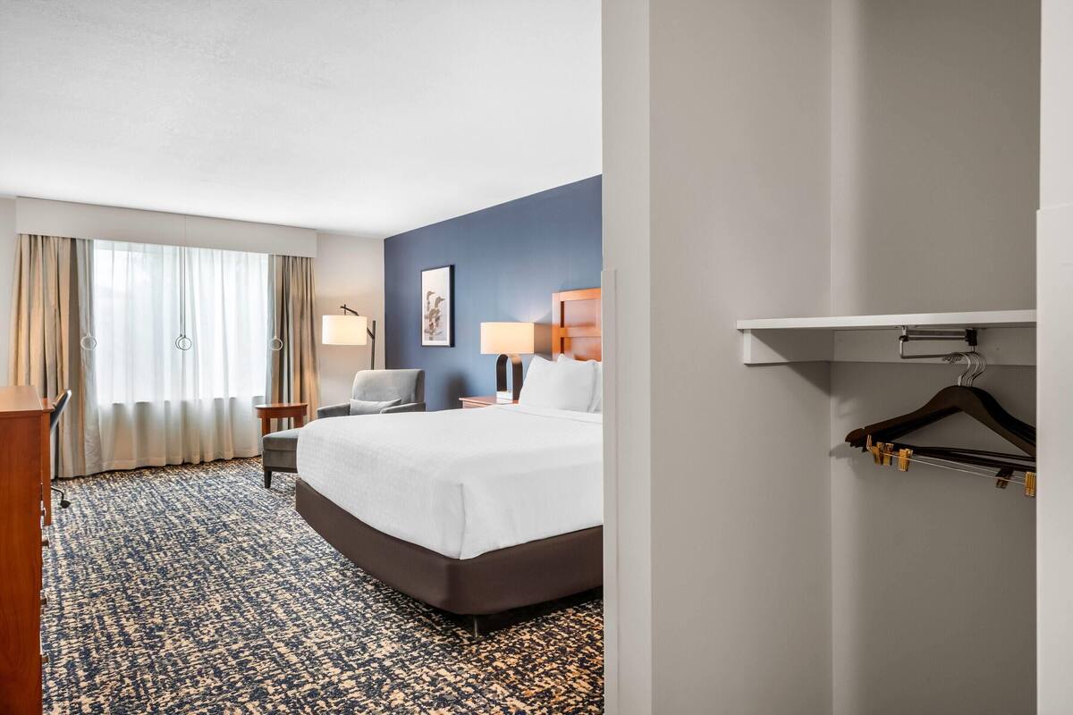 A hotel room with a large bed, a desk, a chair, a lamp, and a coat rack.