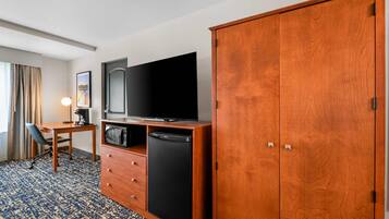 A hotel room with a wooden wardrobe, a flat-screen TV, a microwave, a small refrigerator, a desk with a chair, and a lamp.
