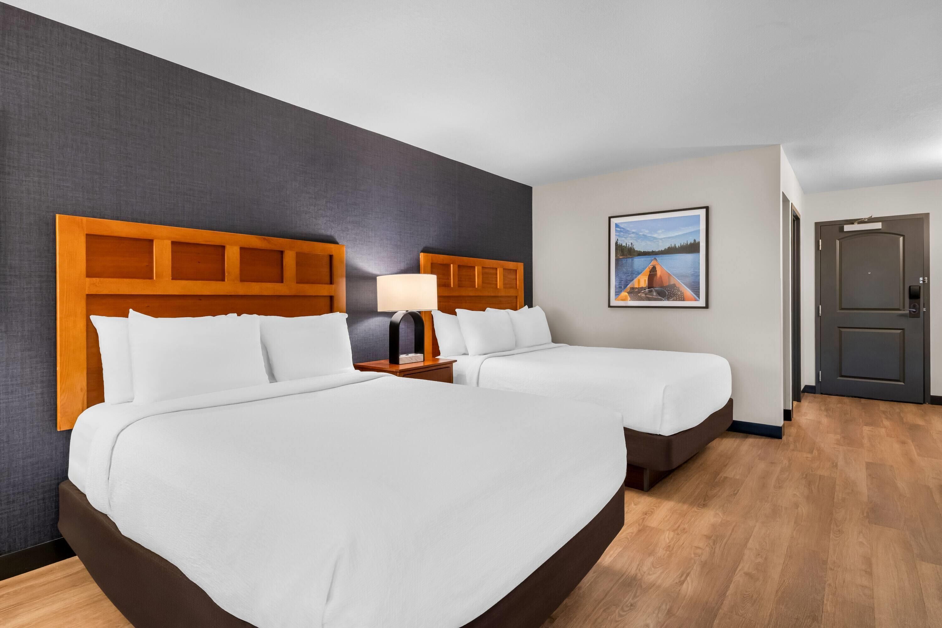 A hotel room with two beds, a nightstand with a lamp, a painting on the wall, and a door.