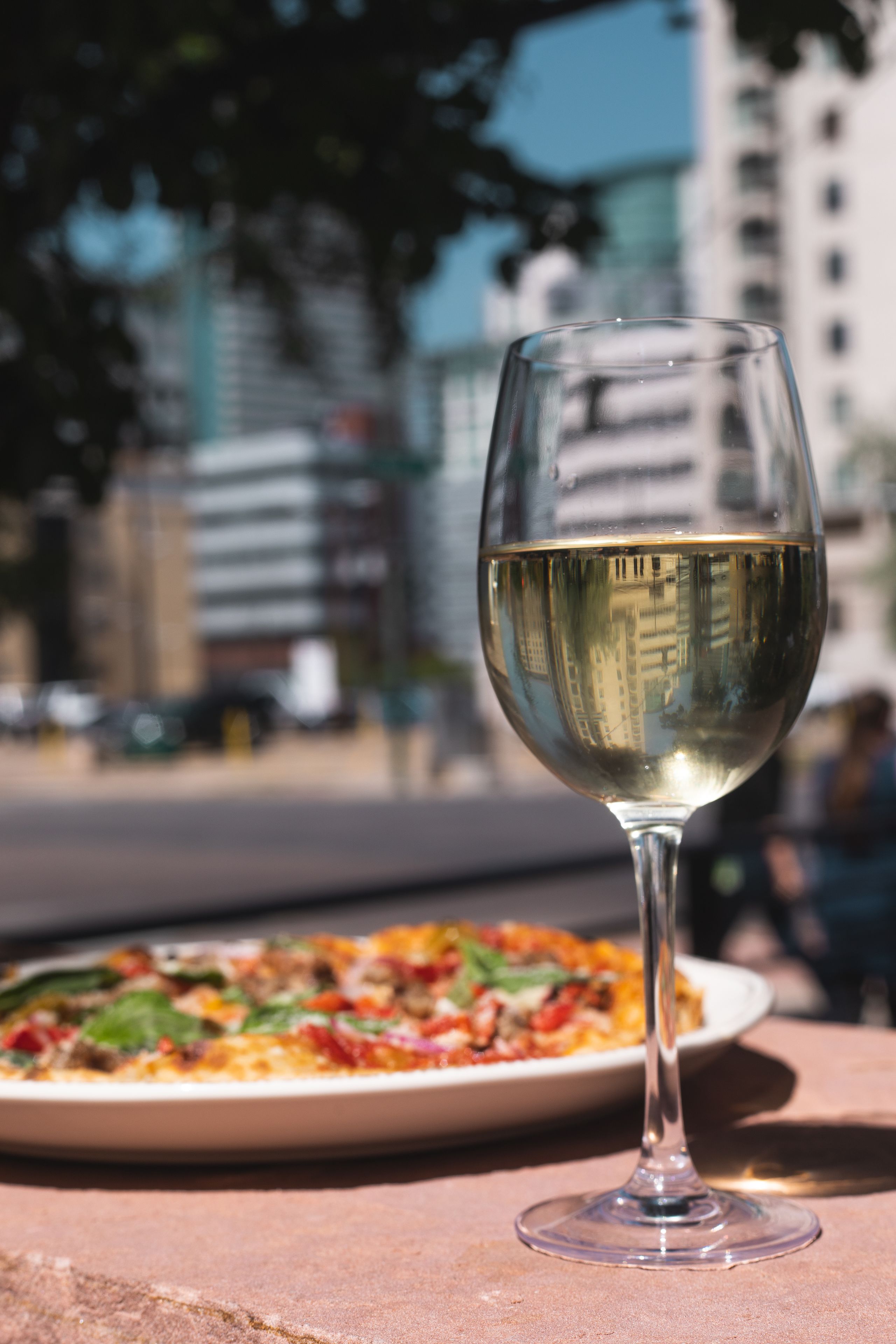A glass of white wine on a table with a pizza in the background.