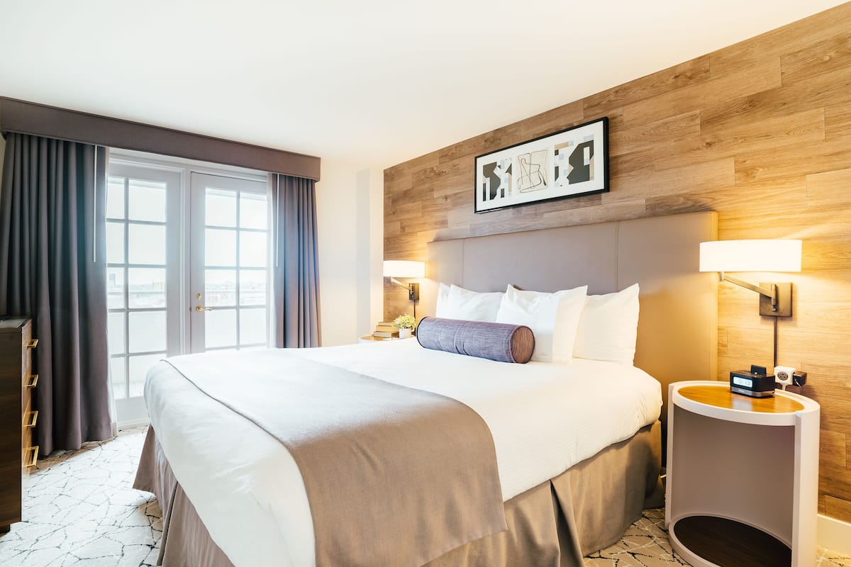 A hotel room with a large bed, a wooden headboard, two bedside lamps, a nightstand, and a view of the city through the window.