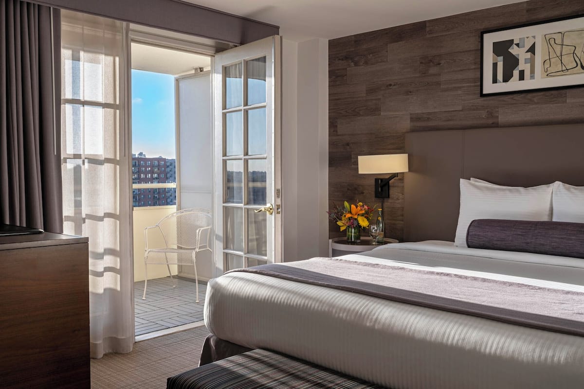 A hotel room with a bed, a desk, a chair, a nightstand with a lamp, and a view of buildings through an open door.