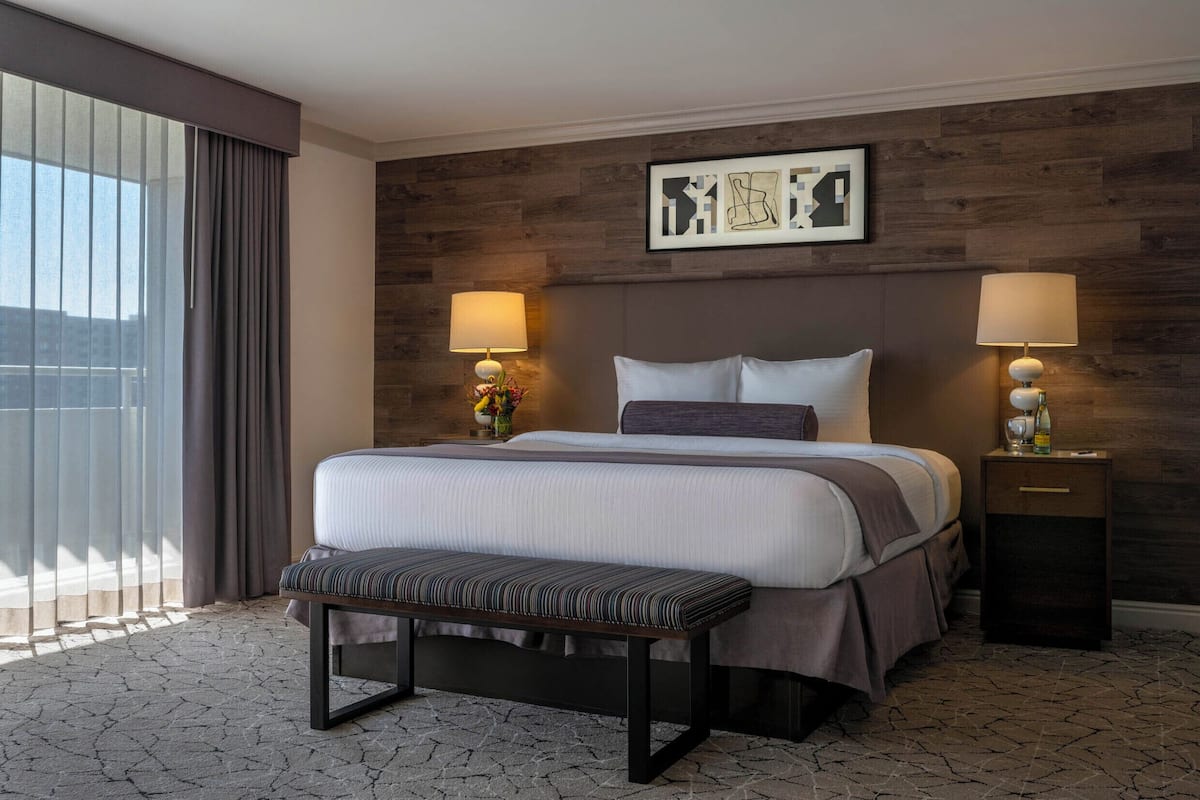A hotel room with a large bed, two bedside lamps, a bench, a wooden headboard, a framed picture, and a window with curtains.