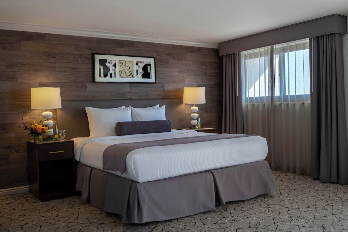 A hotel room with a large bed, two bedside tables with lamps, a framed artwork above the bed, and a window with curtains.