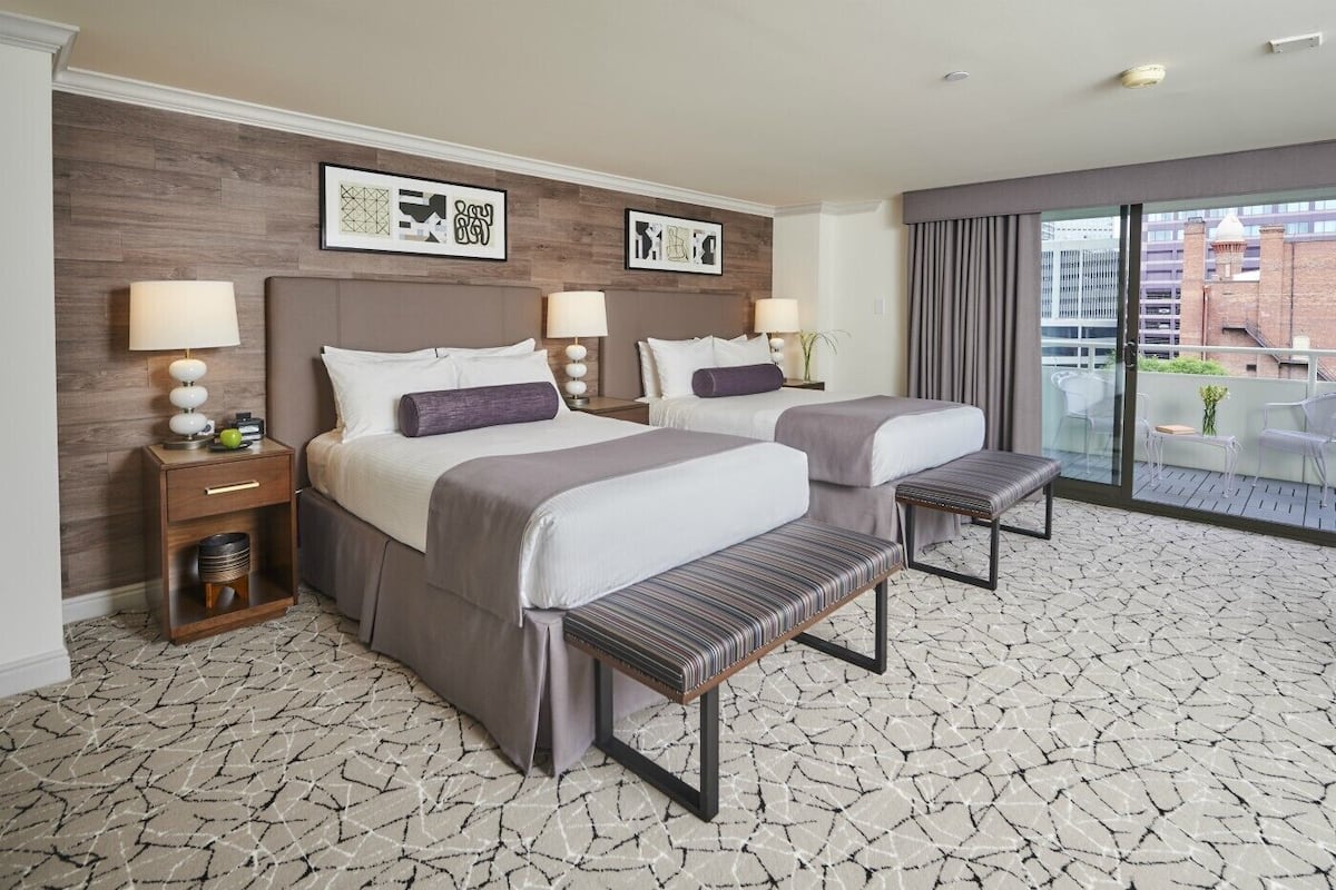 A hotel room with two beds, a small table, a bench, and a balcony with a view of buildings.