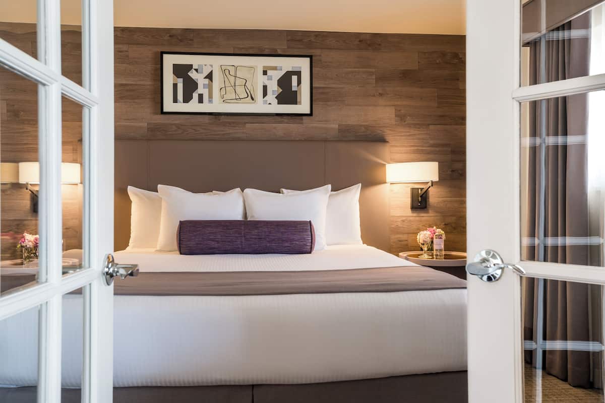A hotel room with a large bed, two bedside lamps, a wooden headboard, and a framed artwork above the bed.