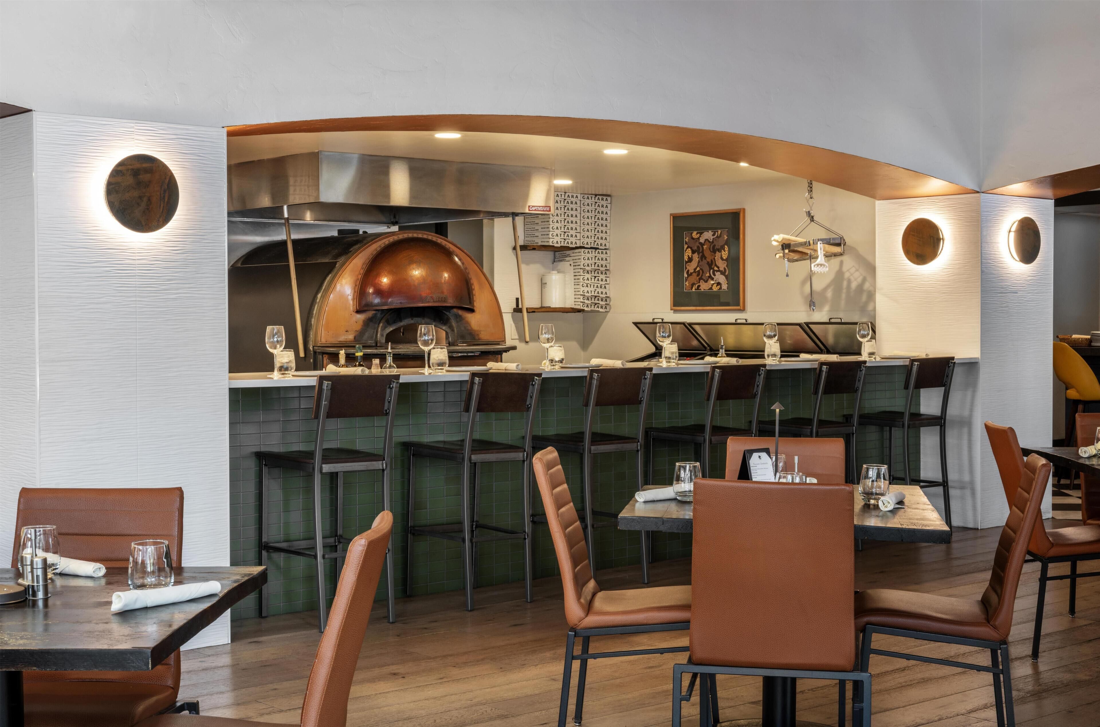 A restaurant interior with a brick oven, high stools, and a menu board.