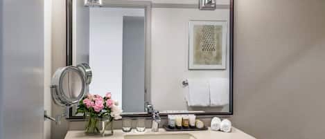 A bathroom with a large mirror, a sink, a round mirror, a vase with flowers, and toiletries.