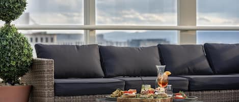 A balcony with a wicker sofa, a potted plant, and a table with food and drinks.