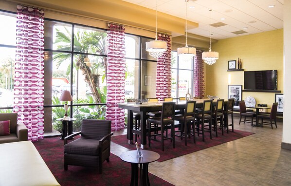 A hotel lobby with a dining area, large windows with curtains, a flat-screen TV, and a view of the outdoors.