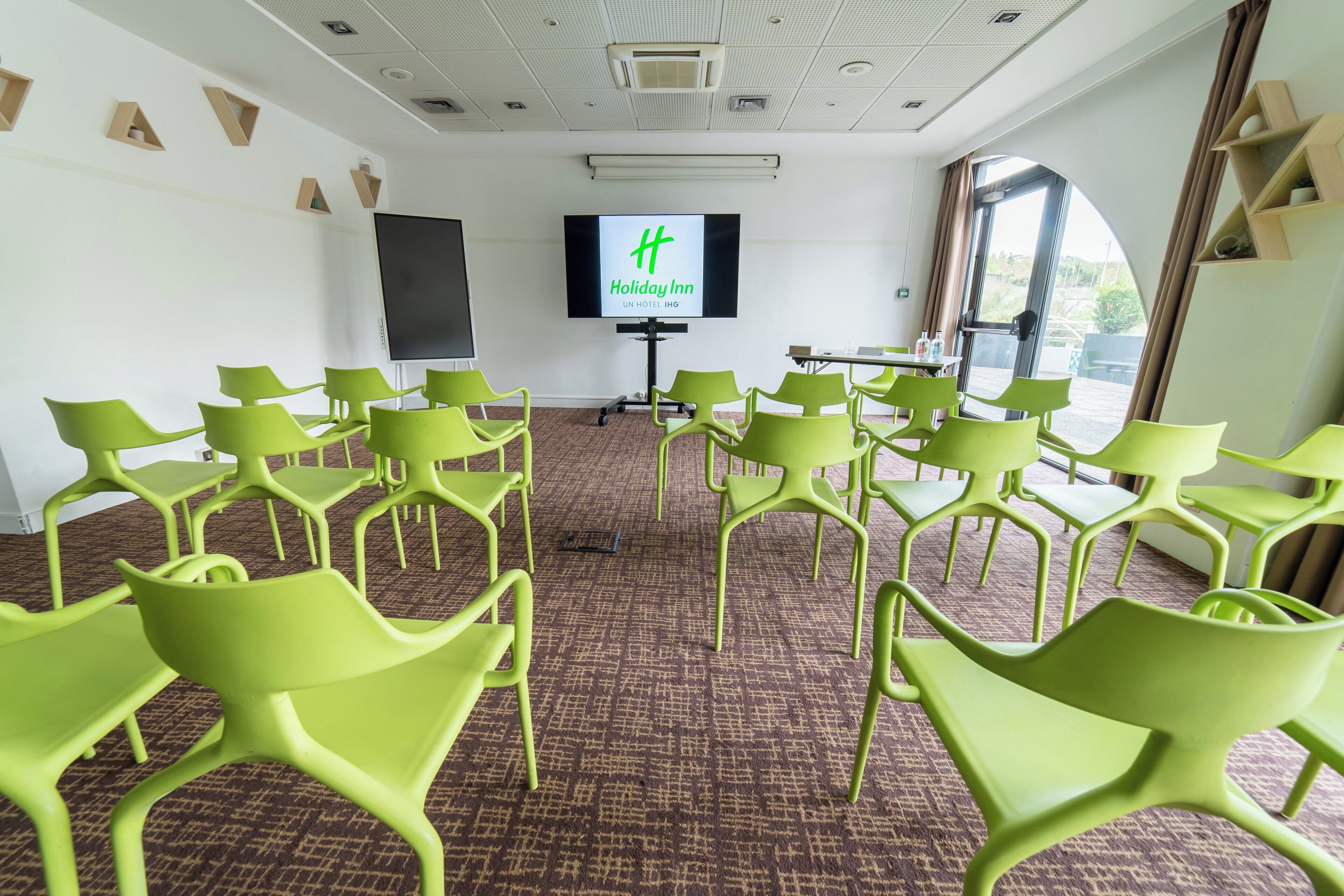Un salón de conferencias con sillas verdes dispuestas en filas, una pantalla grande mostrando el logotipo de Holiday Inn y vistas del exterior a través de un ventanal.
