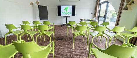 Un salón de conferencias con sillas verdes dispuestas en filas, una pantalla grande mostrando el logotipo de Holiday Inn y vistas del exterior a través de un ventanal.