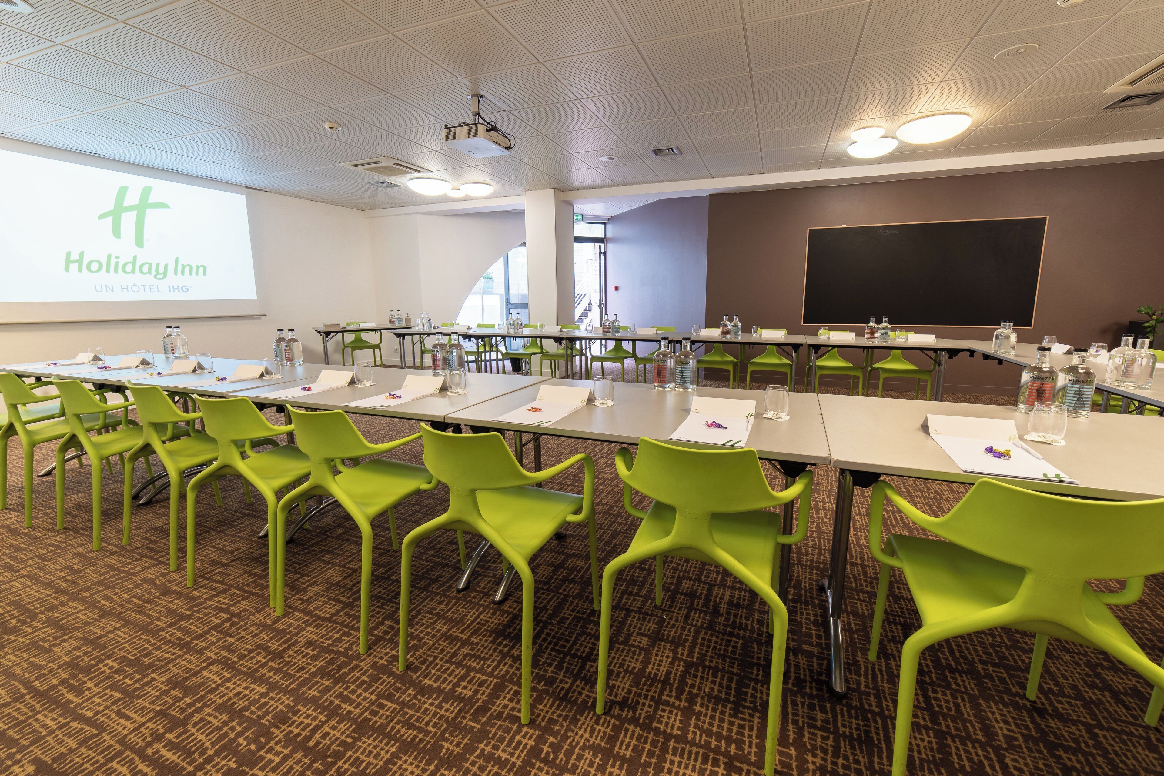 Een vergaderruimte met een lange tafel, groene stoelen, een projectiescherm en een groot whiteboard.