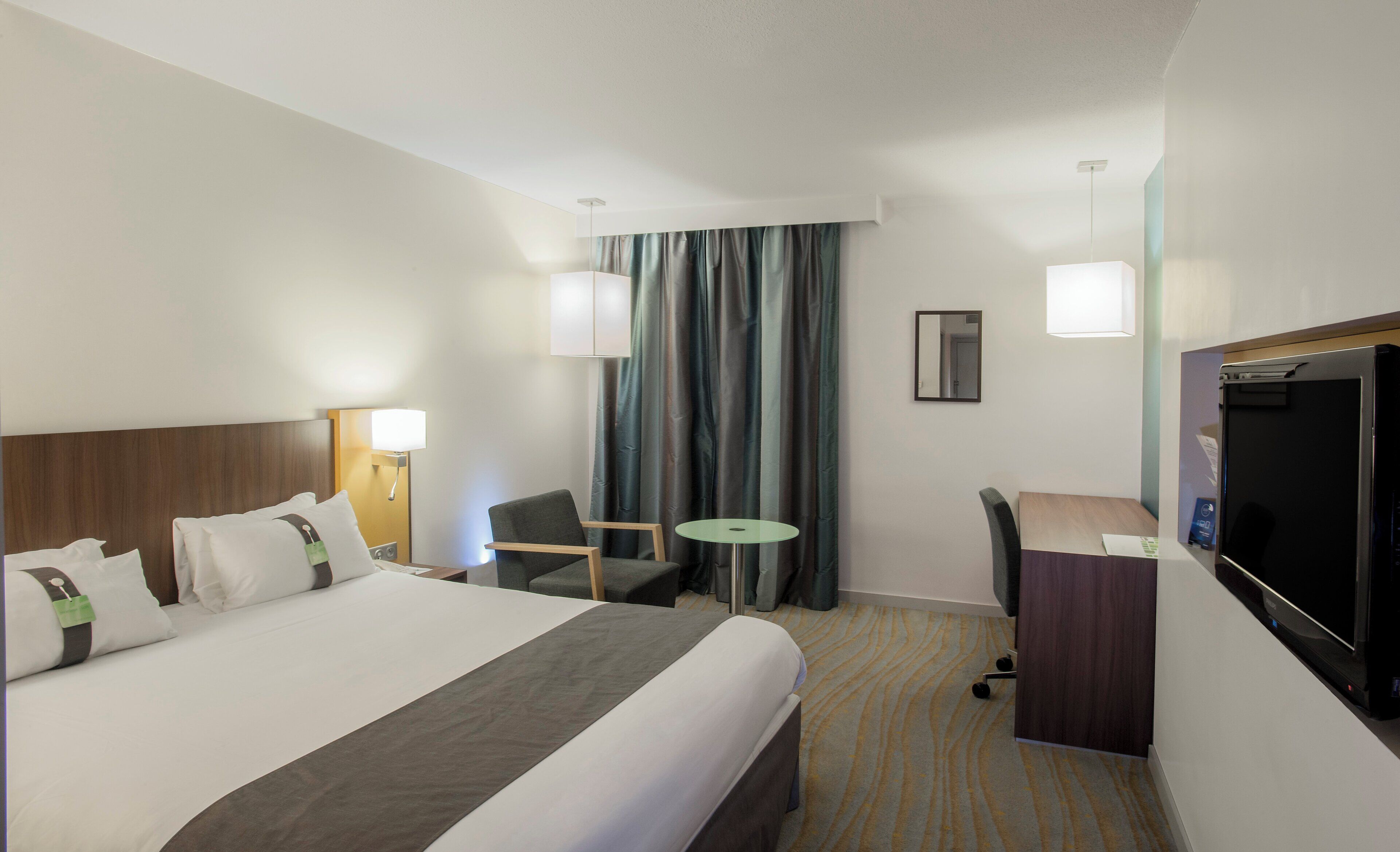 Habitación de hotel con una cama grande, un escritorio, una silla, una mesita, televisión y un espejo.