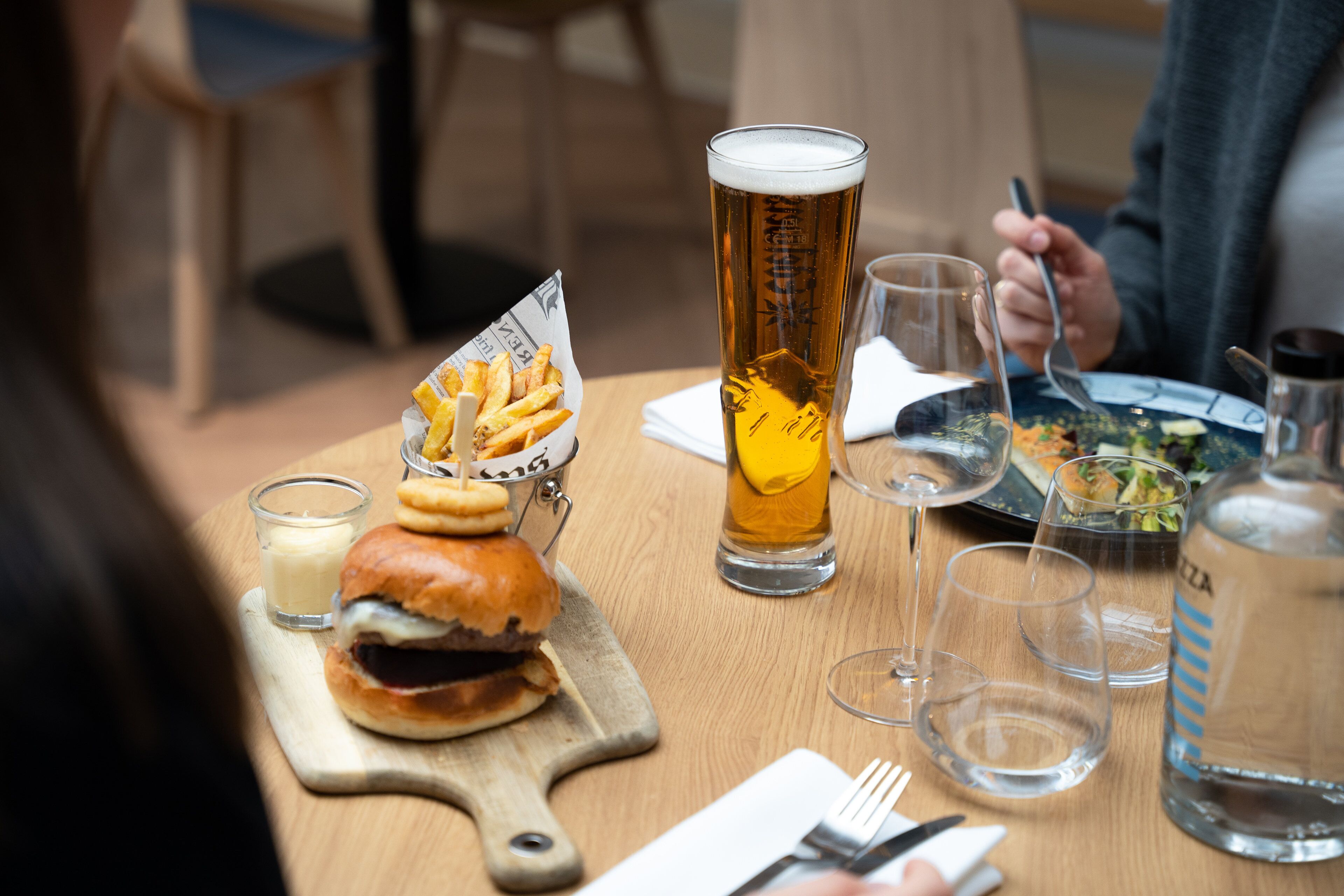 Una mesa de comedor con una hamburguesa, papas fritas, un vaso alto de cerveza y una botella de agua.