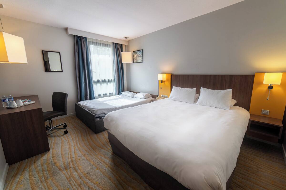 A hotel room with two beds, a desk, a chair, and a large window with curtains.