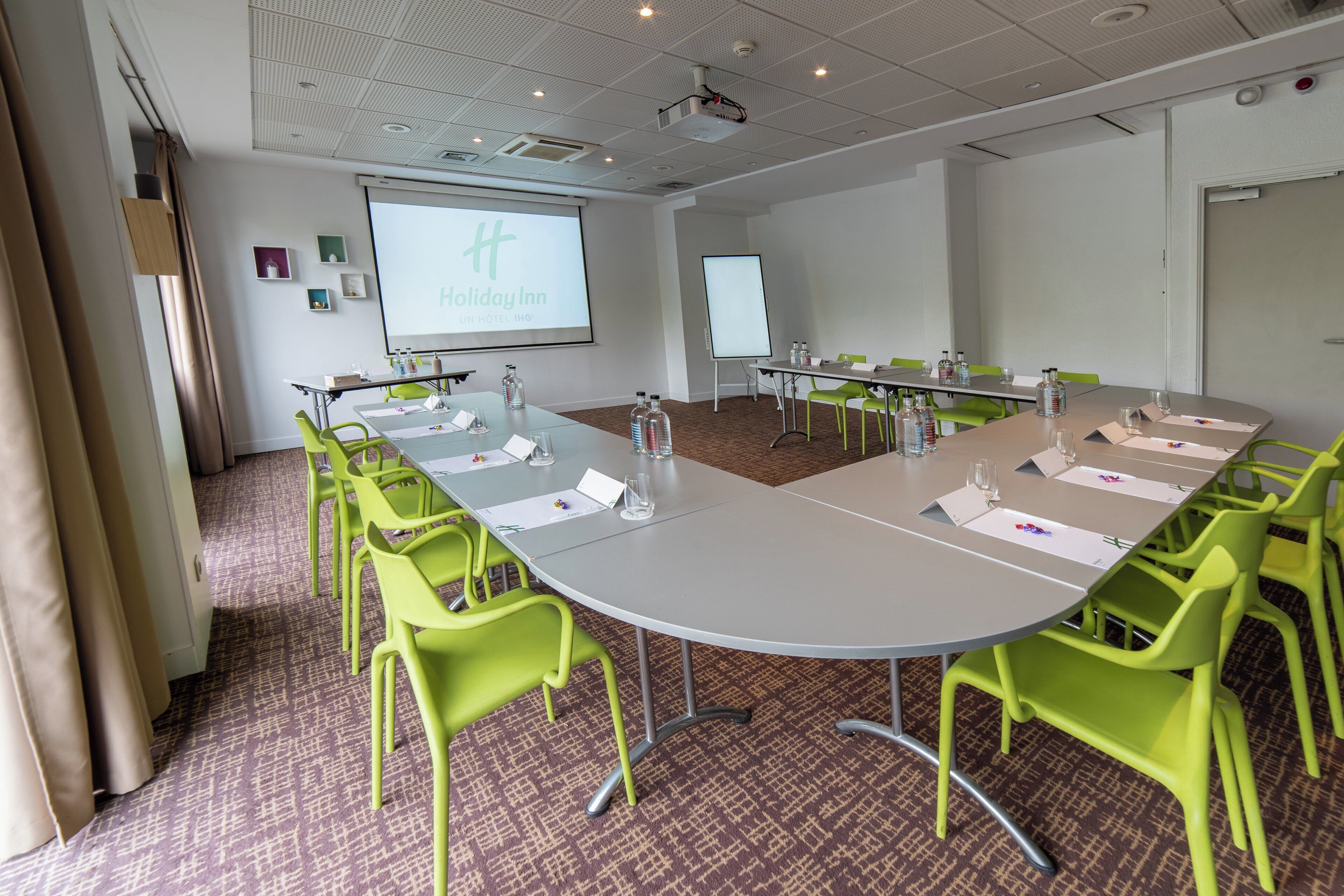 A conference room with a large table, green chairs, and presentation screens.