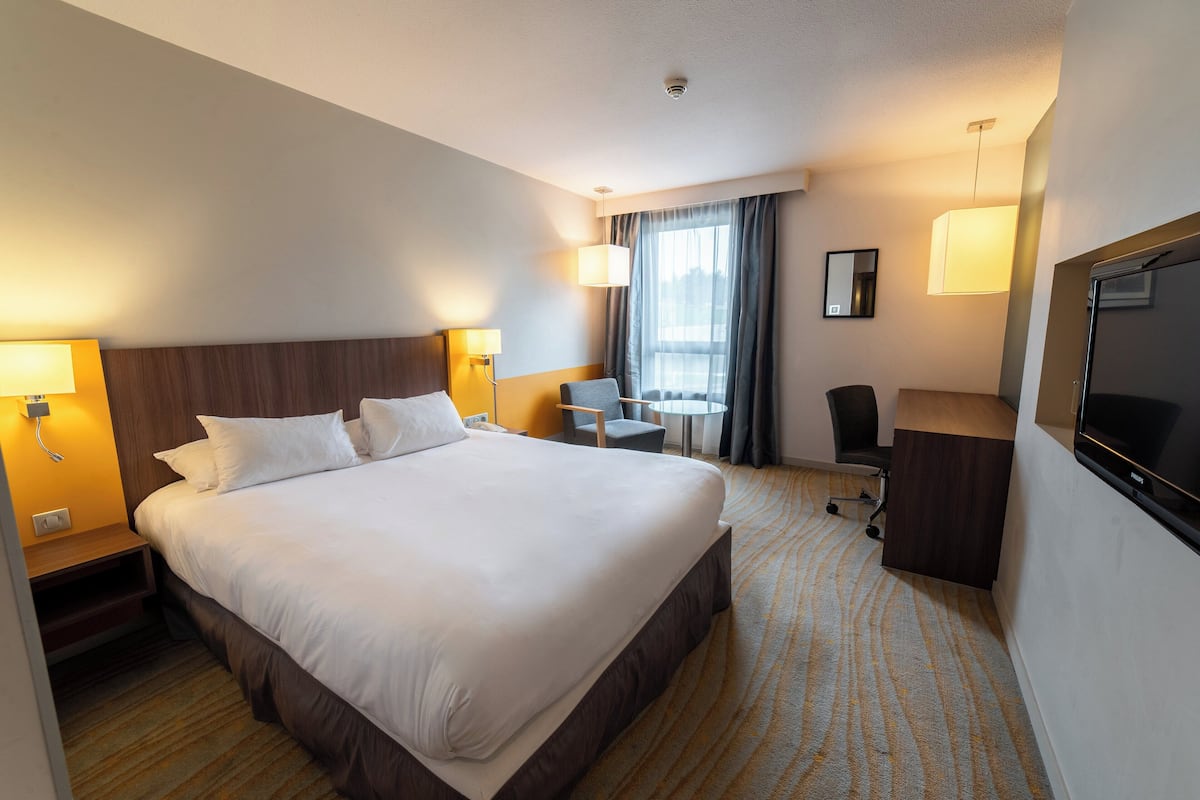 A hotel room with a large bed, bedside tables, a desk, a chair, a TV, and a window with curtains.