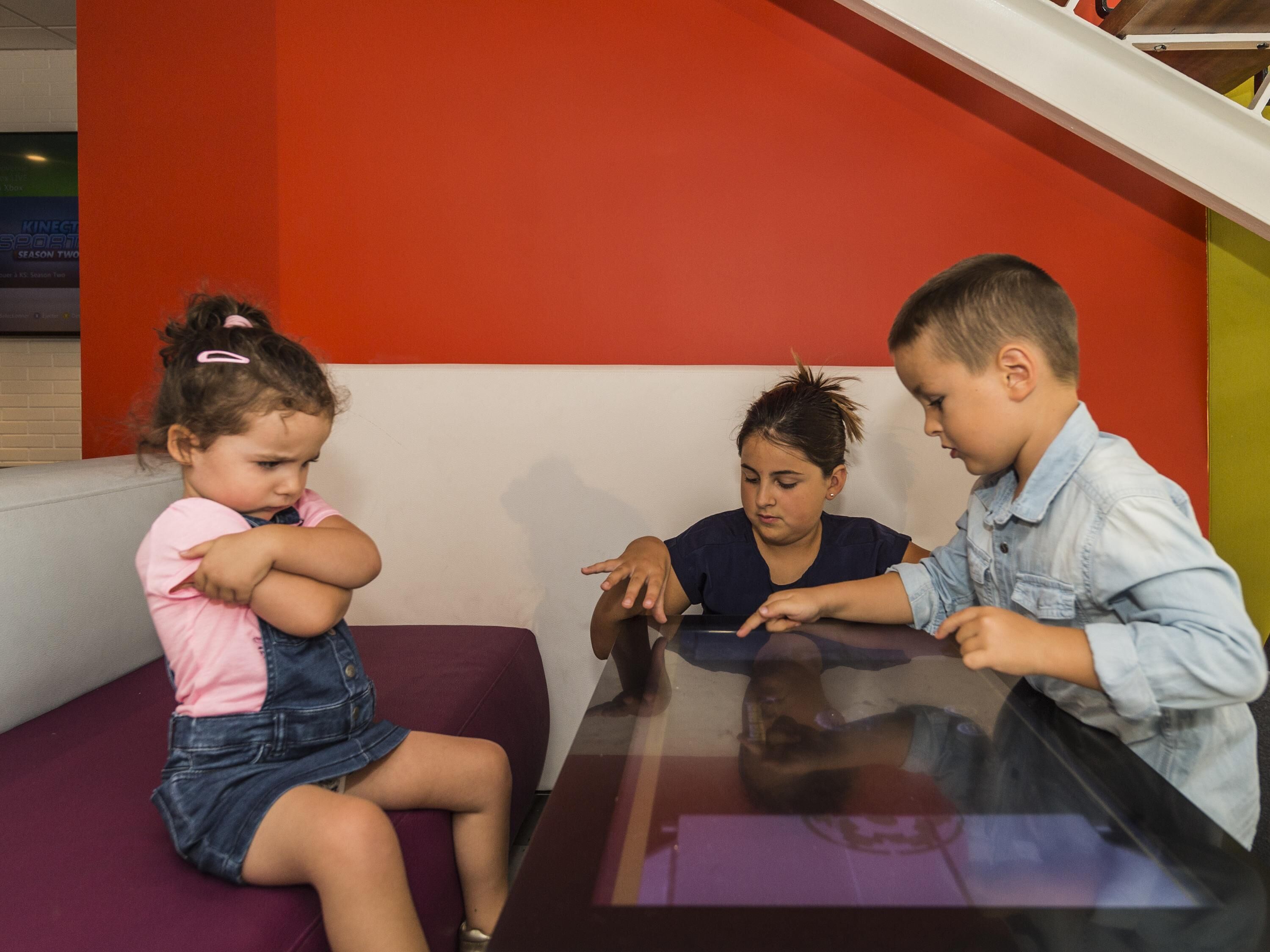 Drie kinderen zitten aan een tafel met een glazen blad, in een kamer met een rode wand en een witte plint.