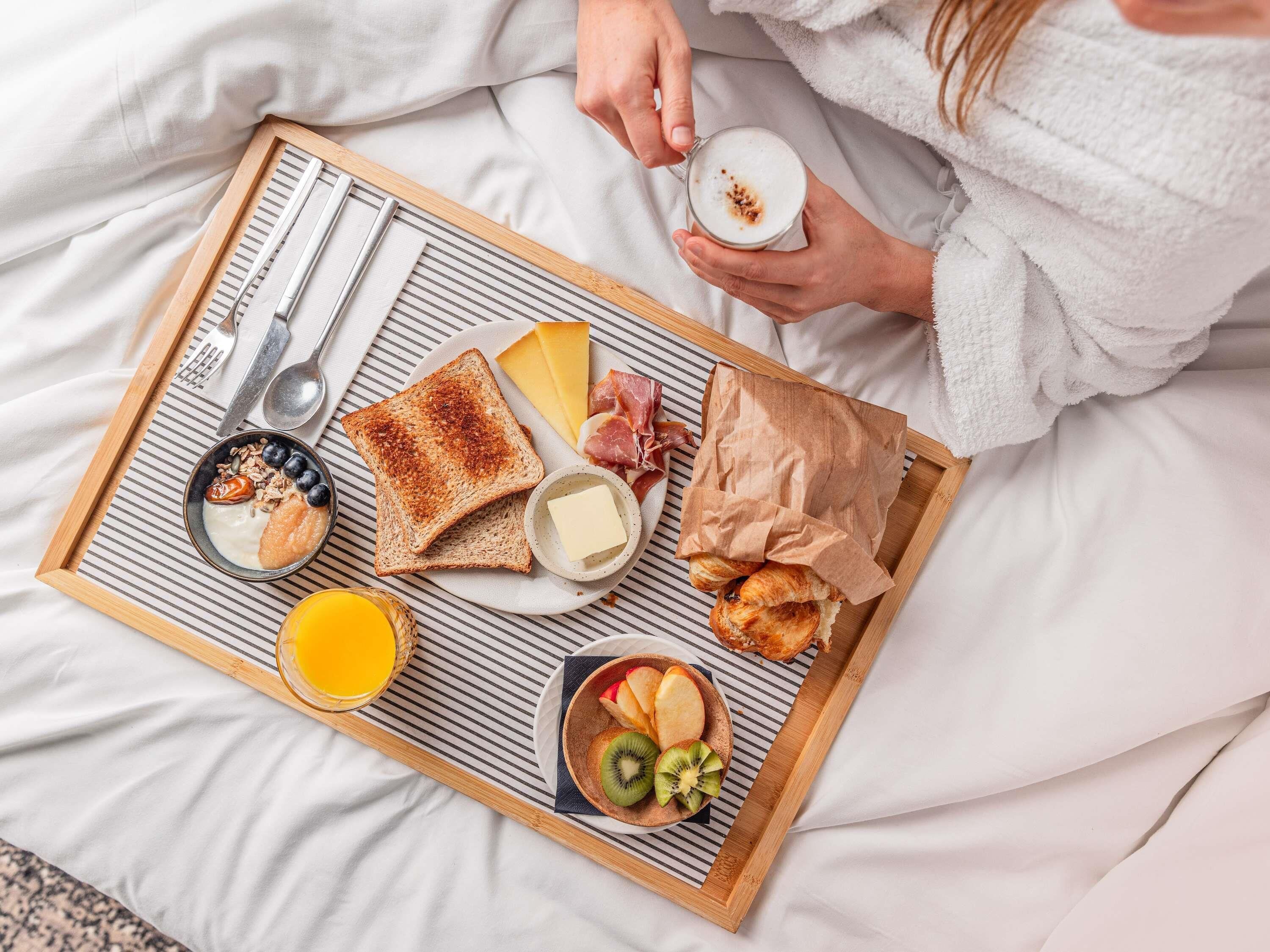 A breakfast tray with toast, cheese, ham, fruit, and a cup of coffee on a bed.