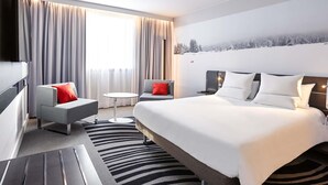 Una habitación de hotel moderna con una cama, dos sillones, una mesa redonda pequeña y un gran ventanal con cortinas.