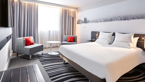 A modern hotel room with a bed, two armchairs, a small round table, and a large window with curtains. - Novotel Lyon Bron Eurexpo (Bron)