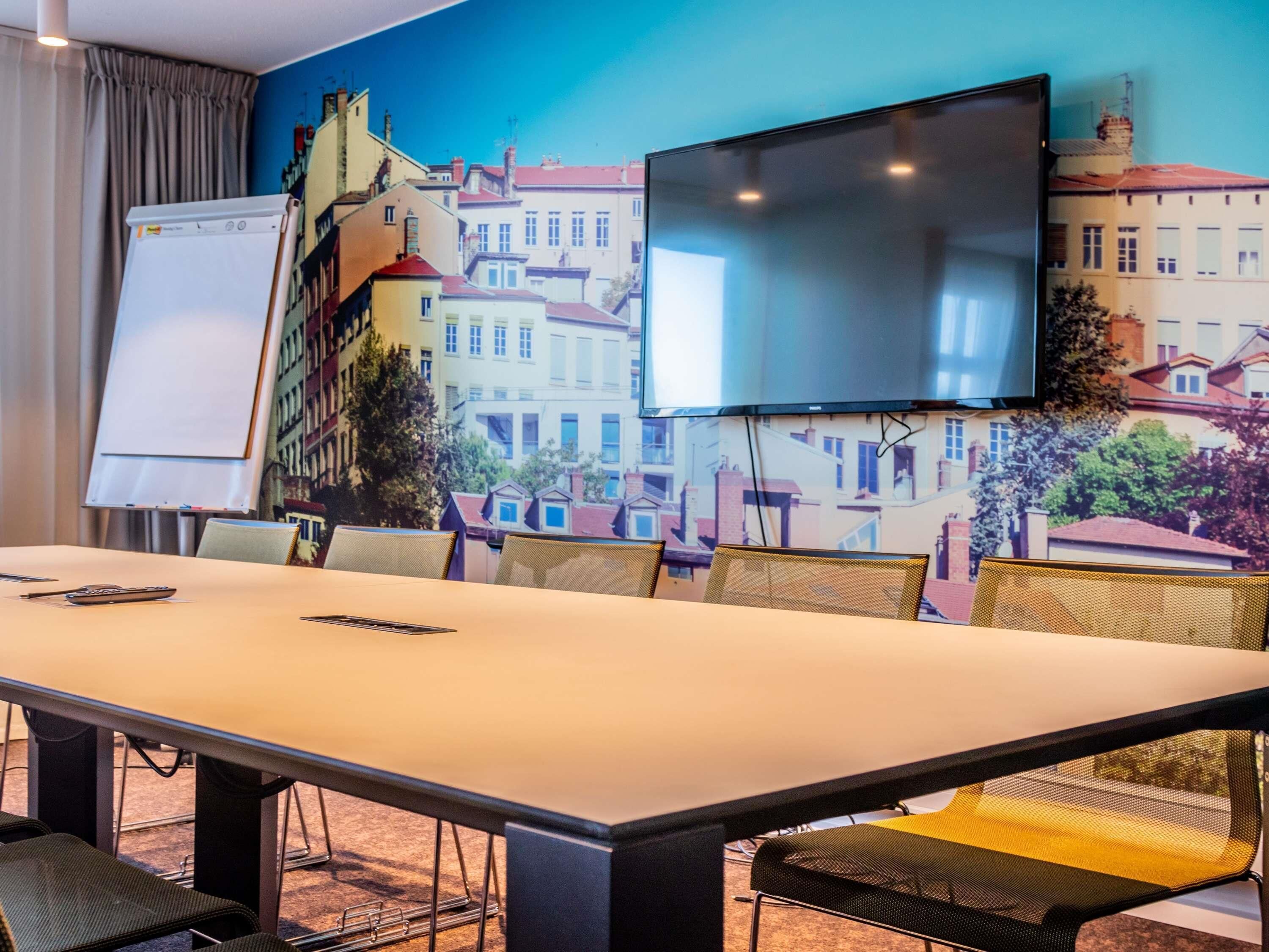 Een vergaderruimte met een lange tafel, stoelen, een whiteboard en een groot flatscreen aan de muur.