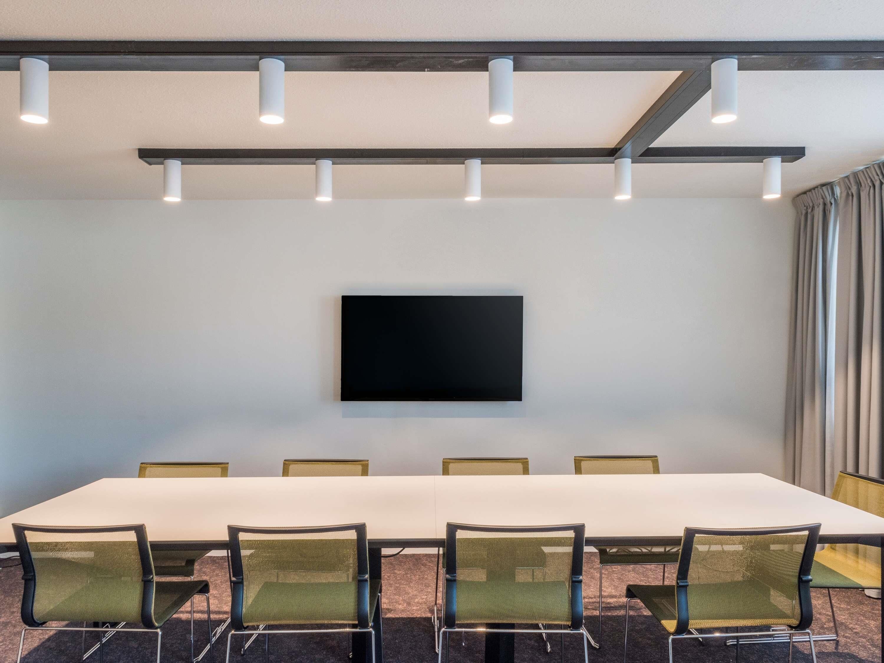 A conference room with a long table, six chairs, a flat-screen TV mounted on the wall, and ceiling lights.