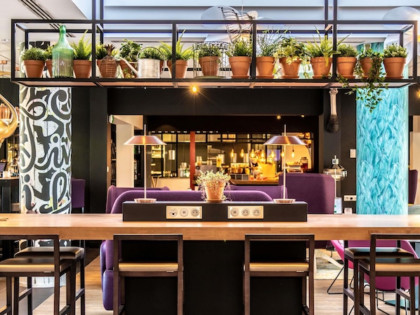 Uma área de bar moderna com balcão de madeira, assentos roxos e plantas em vasos numa prateleira.