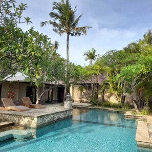A pool area with clear blue water, surrounded by lush greenery and palm trees. There are lounge chairs and a stone wall. - The Royal Beach Seminyak Bali (Seminyak)
