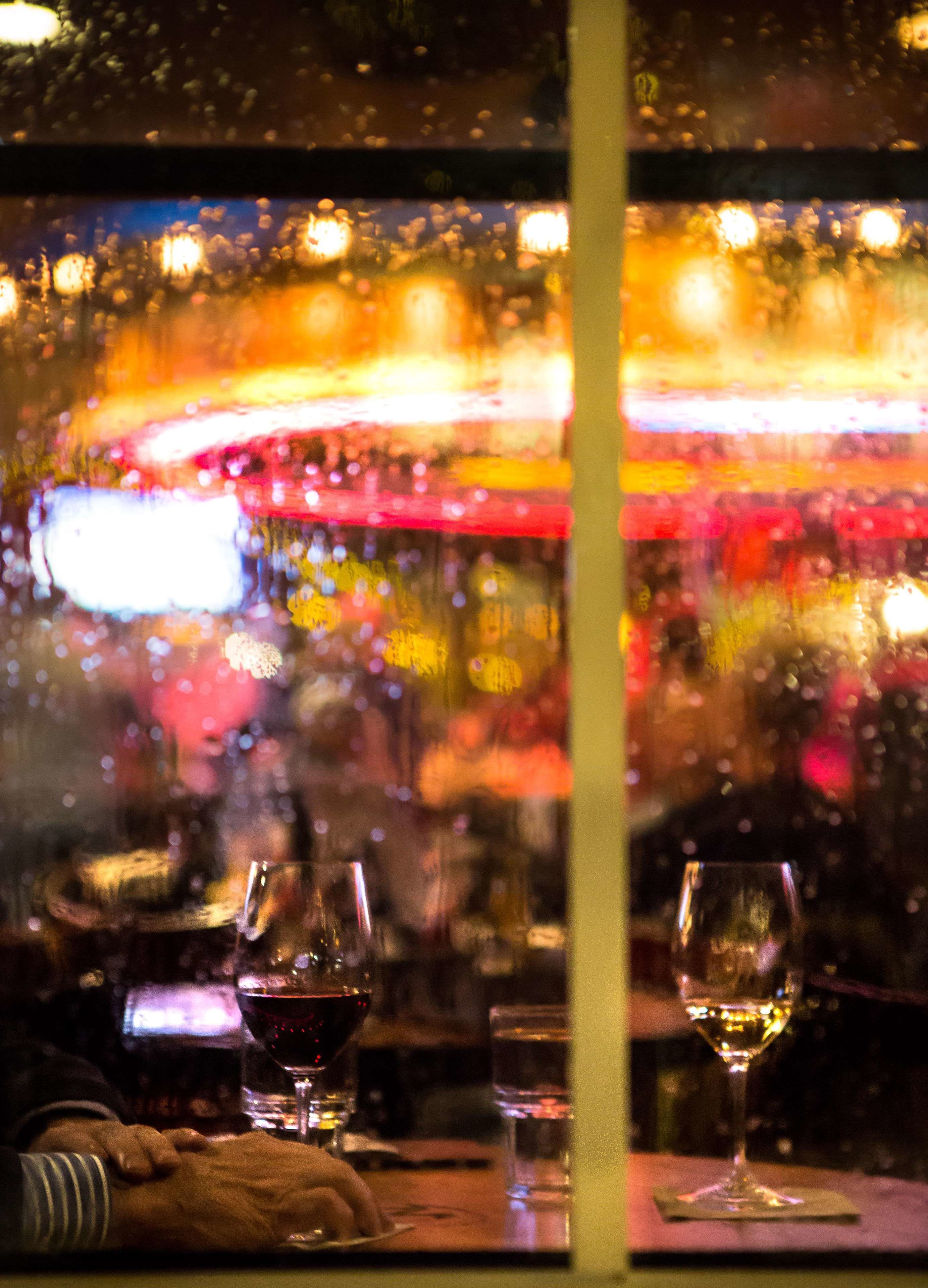 A view through a window with raindrops, two glasses of wine, and a blurred background with lights.