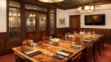 Uma sala de conferências com uma longa mesa de madeira, cadeiras, um candelabro, janelas com vitrais e uma TV de tela plana.