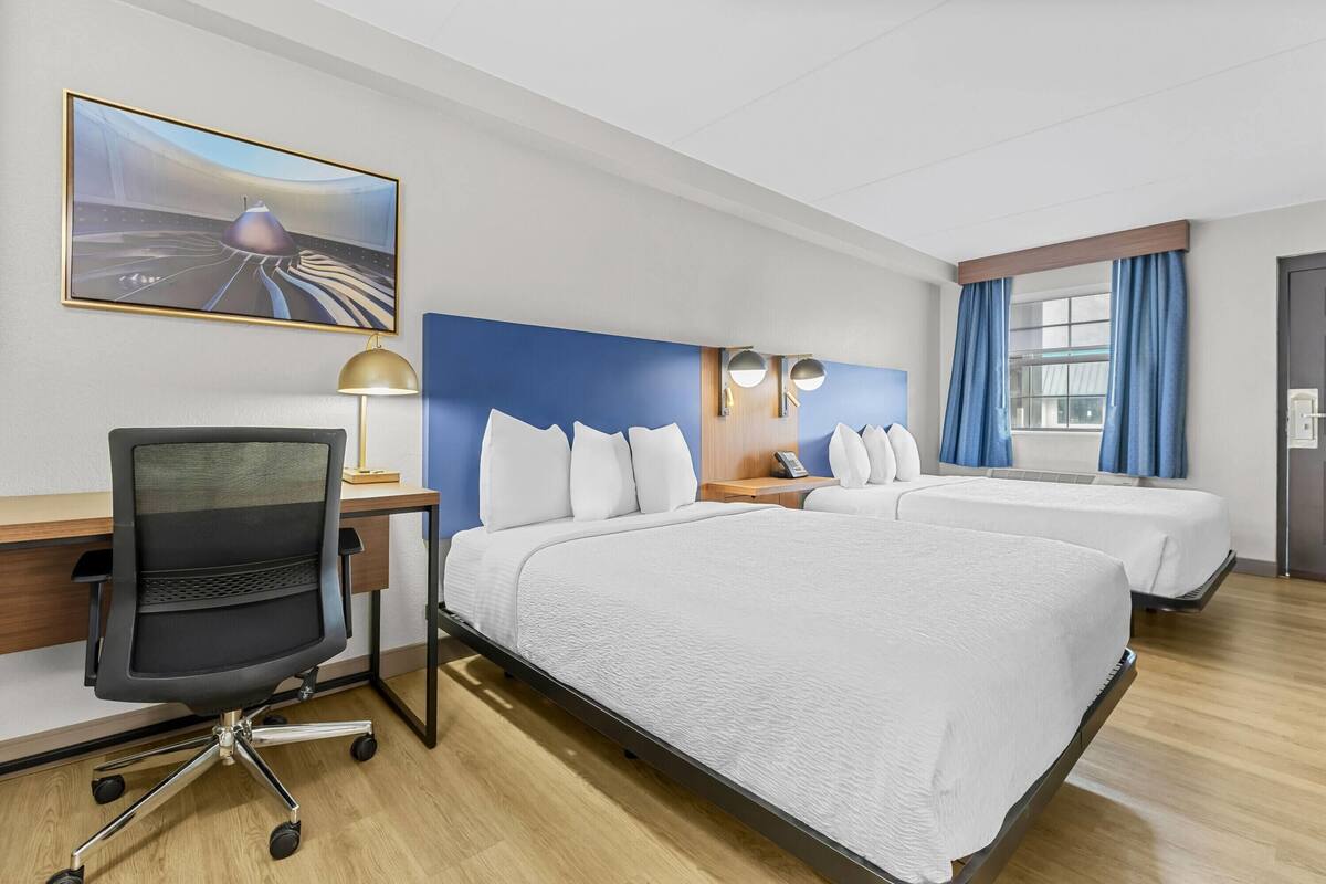 Habitación de hotel con dos camas, un escritorio con silla, una lámpara de escritorio, un cuadro en la pared y una ventana con cortinas.