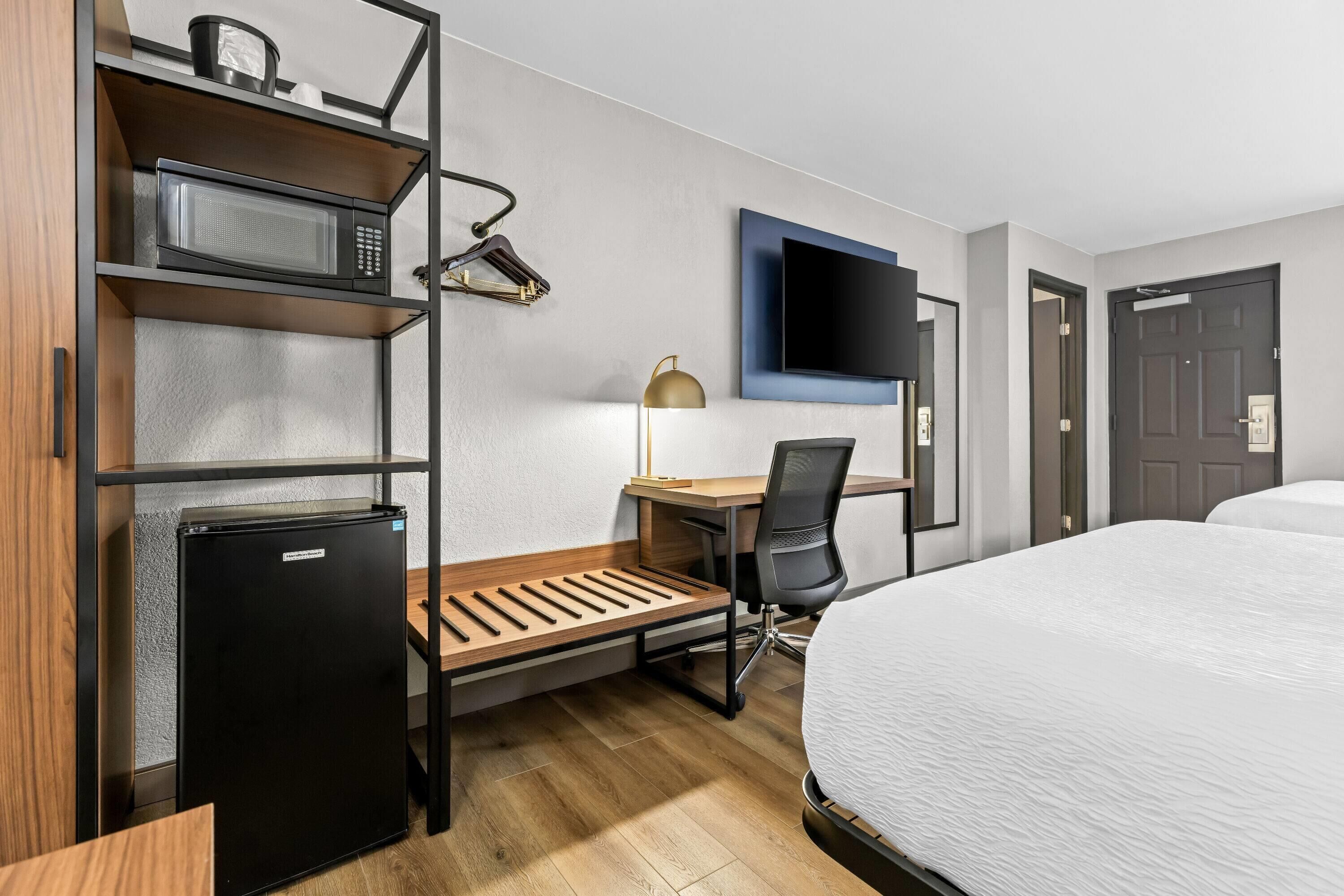 A hotel room with a bed, a desk with a chair, a microwave, a refrigerator, a TV mounted on the wall, and a door.