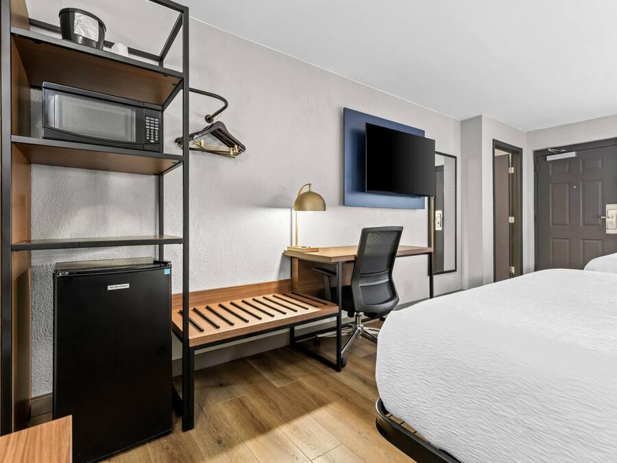 A hotel room with a bed, a desk with a chair, a microwave, a refrigerator, a TV mounted on the wall, and a door.