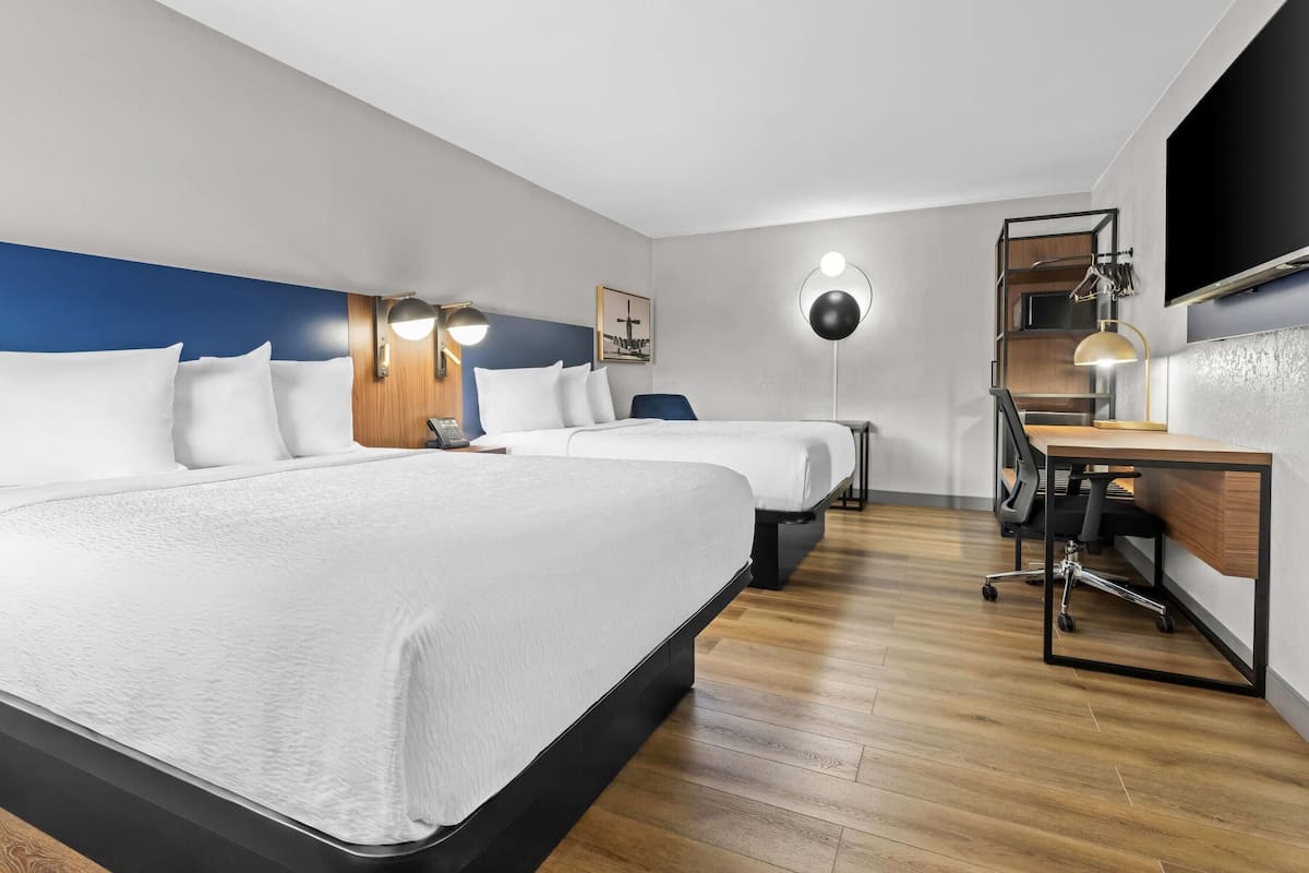 Habitación de hotel con dos camas, un escritorio, una silla, un televisor y un estante en la pared.