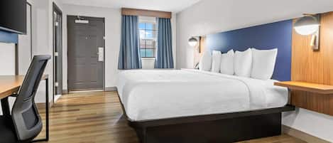 A hotel room with a bed, desk, chair, and a view of the outdoors through a window.