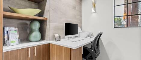 Un espace de bureau moderne avec un bureau blanc, un ordinateur et une chaise. On y trouve des étagères en bois ornées d’objets décoratifs et une fenêtre donnant sur l’extérieur.