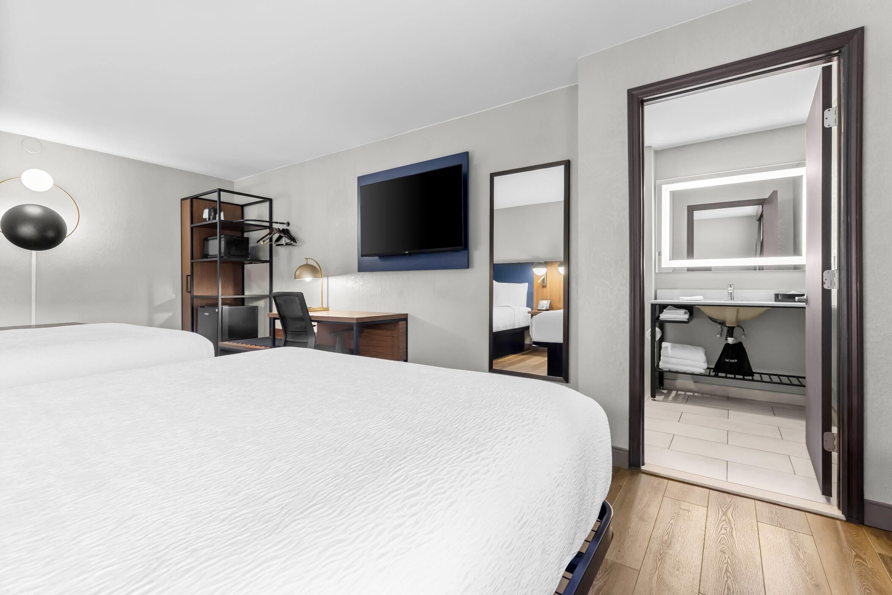 Habitación de hotel con una cama grande, un escritorio con una silla, un televisor montado en la pared, un armario con espejo y una lámpara de noche.