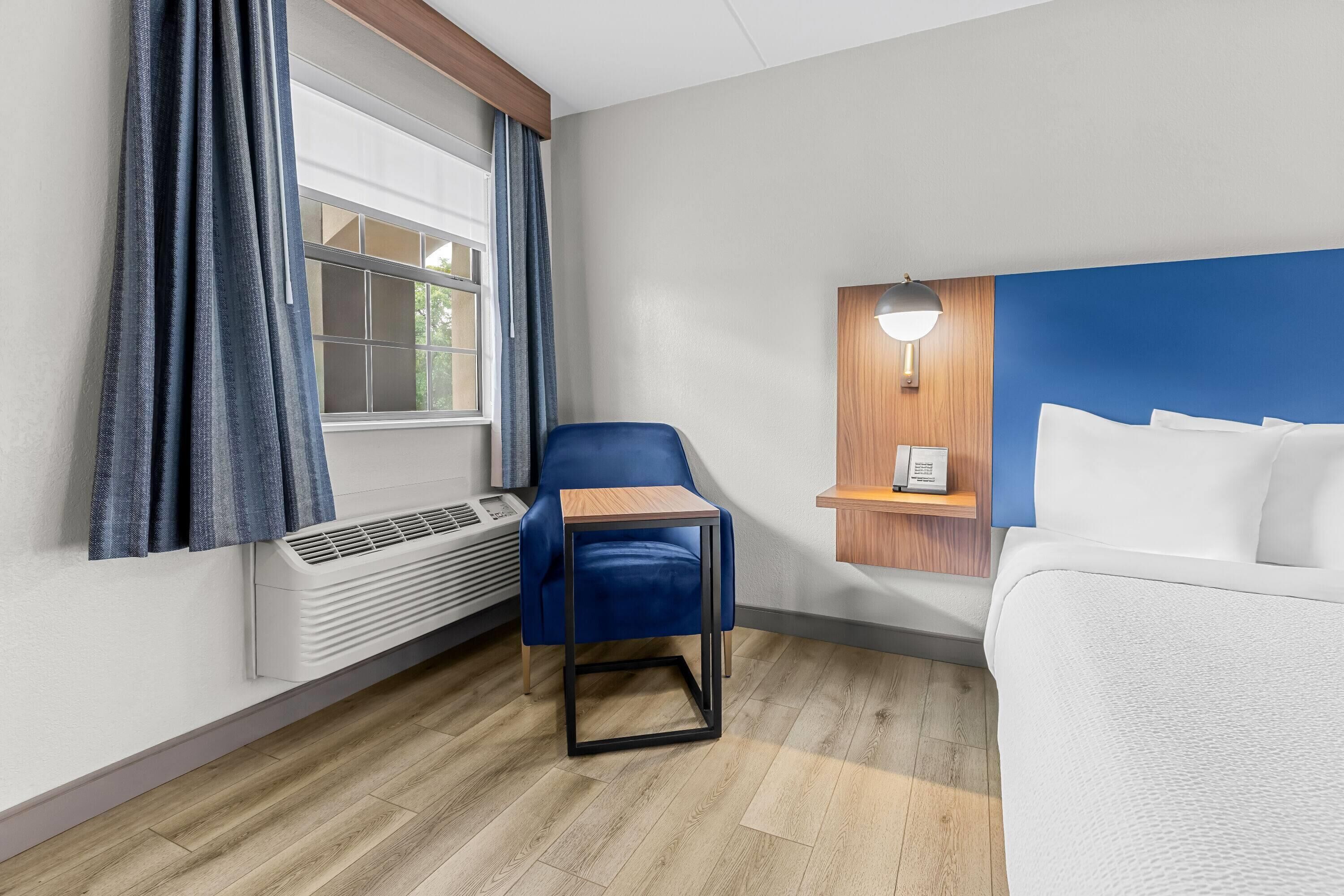 Ein Hotelzimmer mit einem Bett, einem blauen Sessel, einem kleinen Tisch, einer Wandablage und einem Fenster mit Vorhängen.