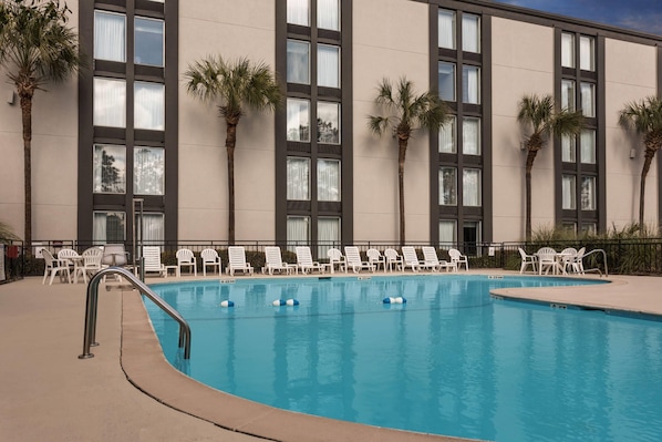 A swimming pool with lounge chairs and palm trees, surrounded by a building with large windows and a beige facade.
